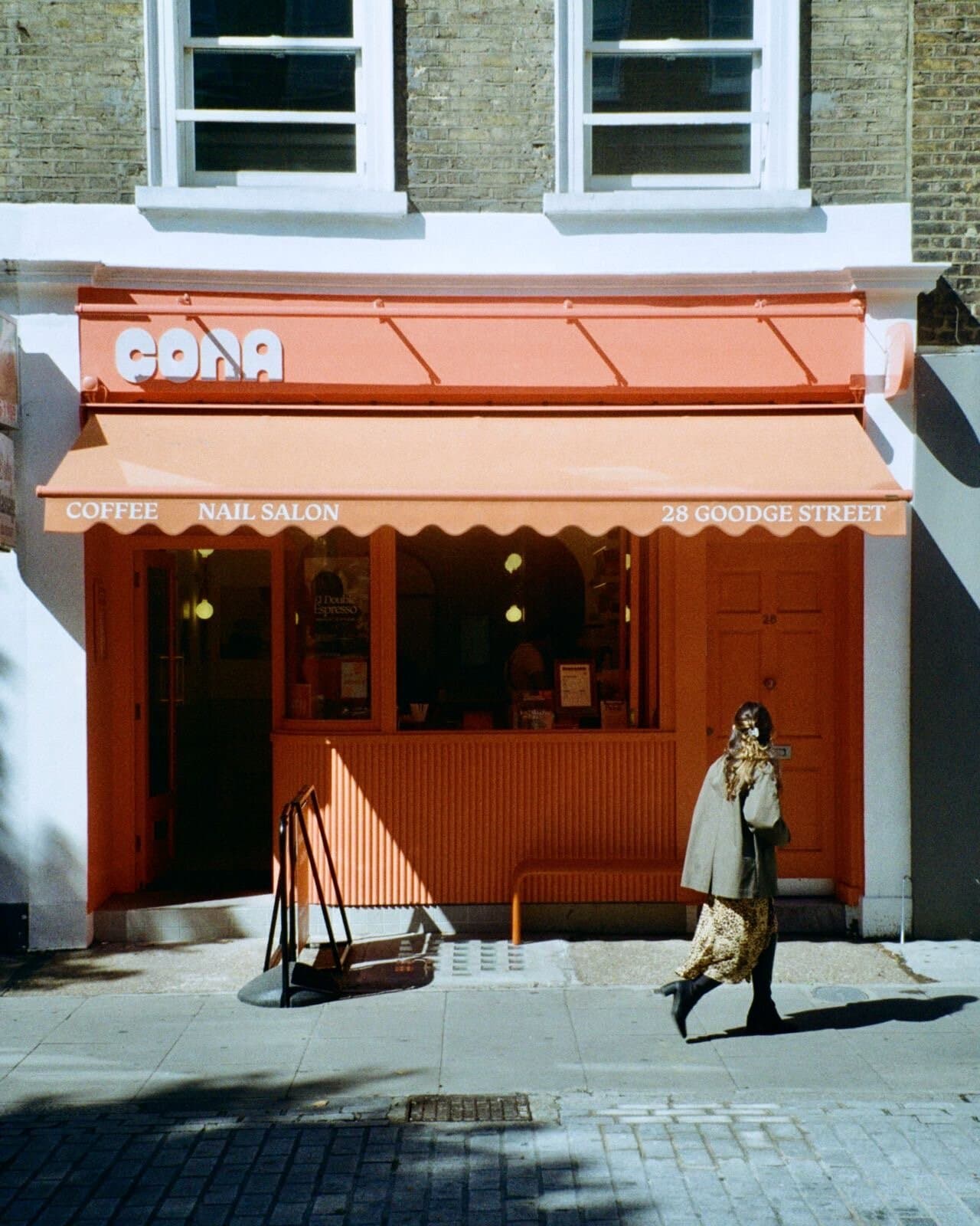 The CONA storefront on 28 Goodge Street — terracotta awning reading COFFEE / NAIL SALON, with a passer-by in the afternoon sun